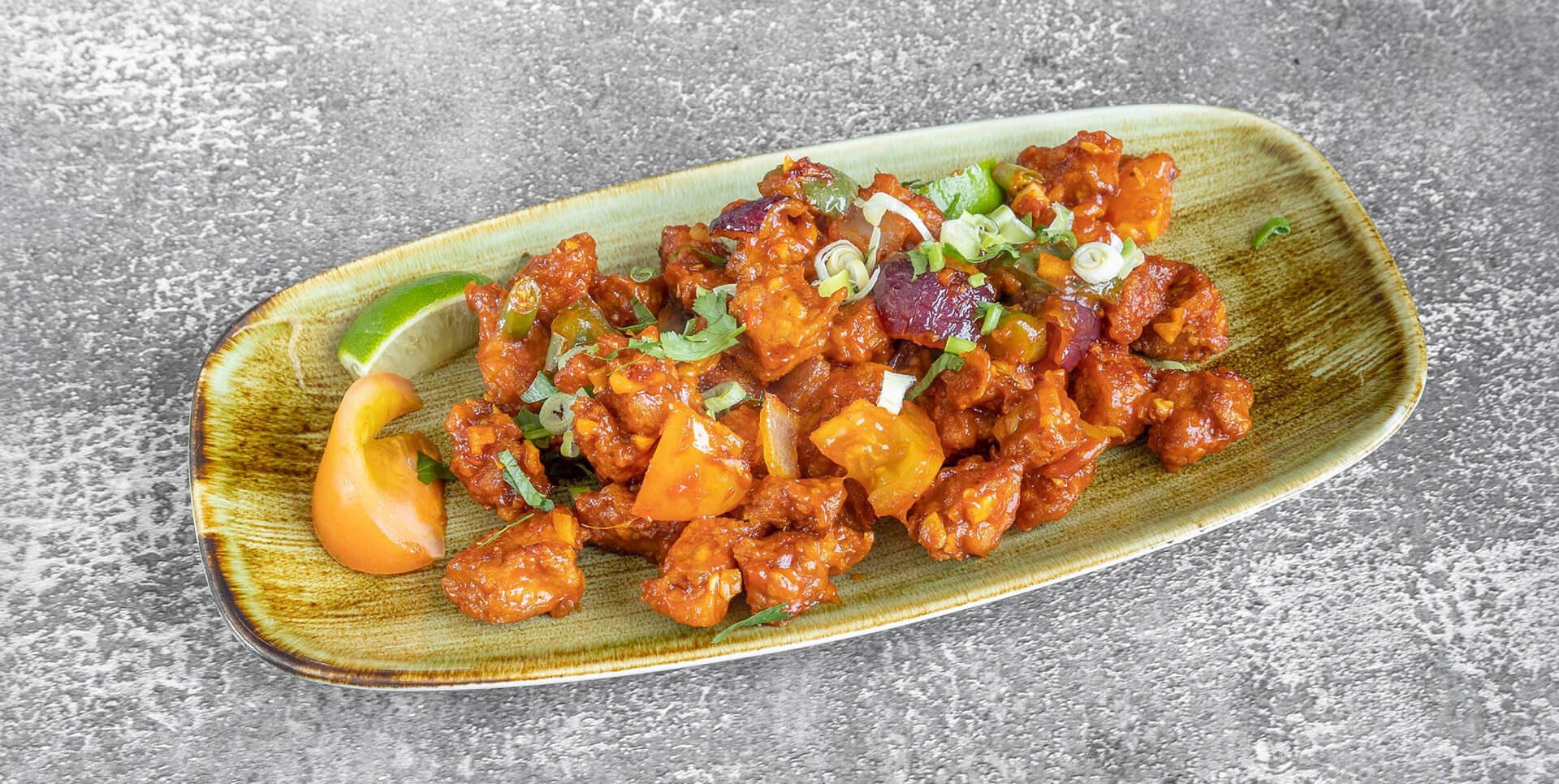 Spicy fried chicken dish garnished with sliced spring onions and cilantro. Served on a rectangular dish with a lime wedge and tomato slice, atop a textured surface.