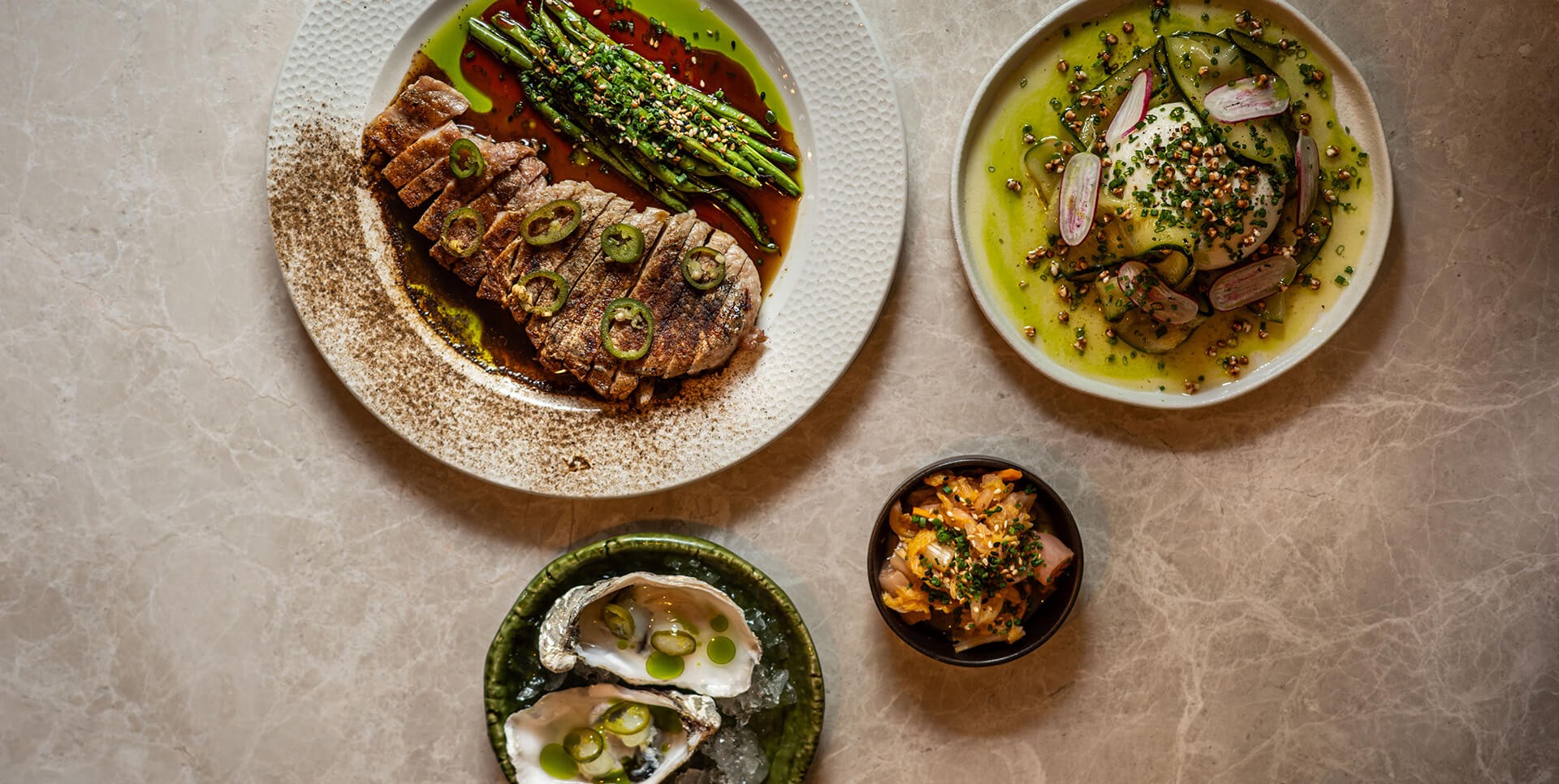 A variety of dishes including a sliced steak with jalapeño slices and a side of glazed asparagus, a plate of burrata with thinly sliced cucumbers and radishes in a vibrant green sauce, fresh oysters on ice with herb oil, and a small bowl of kimchi-style pickled vegetables