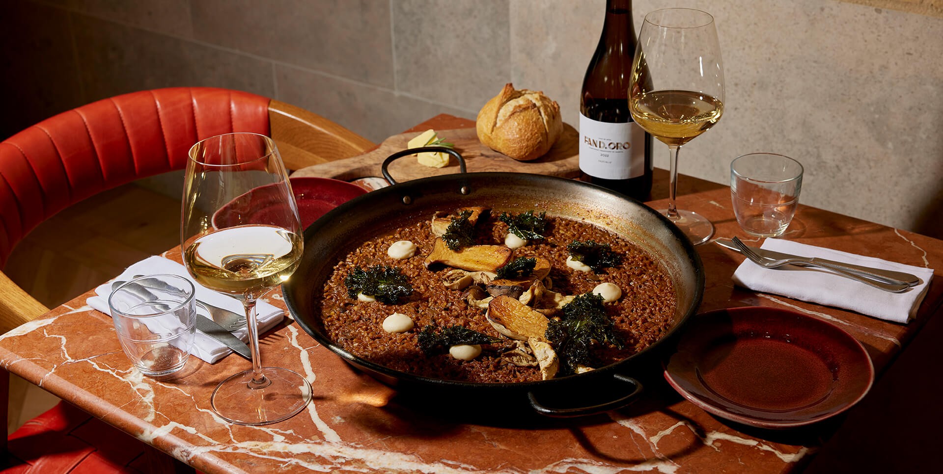 A set dining table displaying a large pan of Spanish risotto topped with wild mushrooms, crusty bread roll and two glasses of white wine